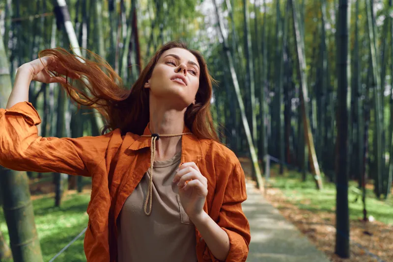 Woman portrait in bamboo forest on a shaded path, fashion movement captured as hair flips and sunlight through foliage, stylish orange jacket and casual pose conveying joy and freedom.