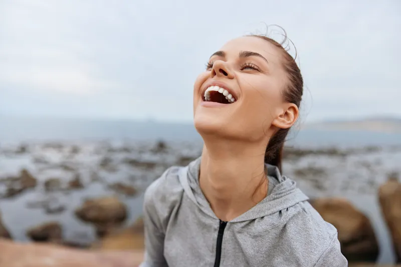 Joyful young woman in a gray sports hoodie laughing outdoors by the sea, expressing happiness and freedom against a rocky landscape backdrop