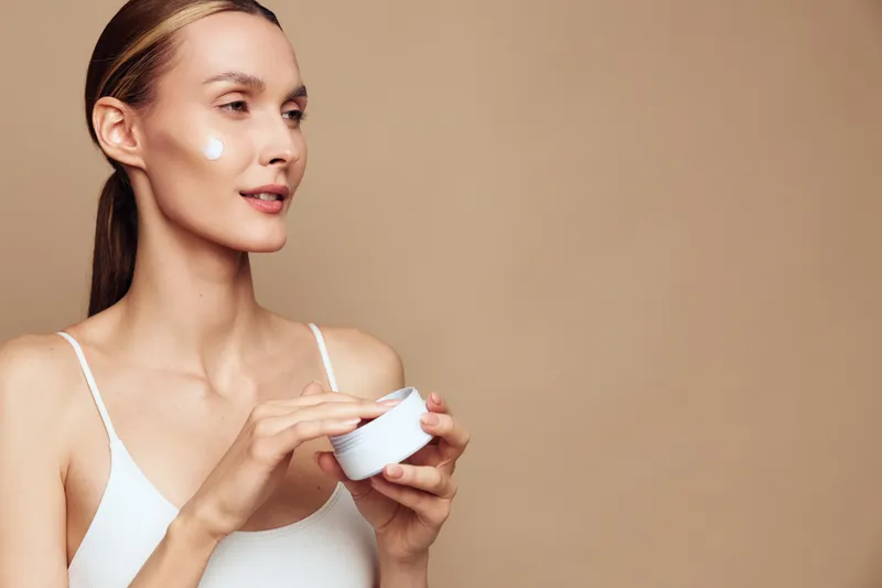 Young woman in a beauty portrait applying face cream from a small jar, smooth hydration skincare concept, calm smile, clean skin care routine, studio beige background, soft natural light
