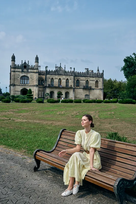 A woman in a light dress sits on a wooden bench in a sunlit park, with a historic castle in the background, creating a peaceful outdoor scene full of calm and timeless charm