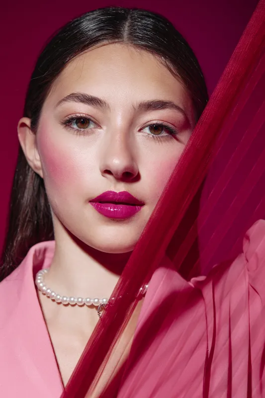 Creative fashion close-up of a young woman with glossy magenta lips, dewy skin, and dramatic blush, wearing light pink clothing and pearls against a vivid pink background.