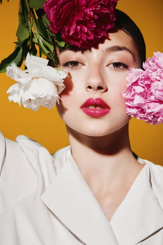Young woman with bright floral makeup holds pink and white peonies near her face against a yellow backdrop. Fashion beauty close-up with creative vibrant styling and glossy lips.