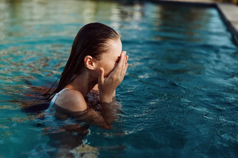Candid woman in an outdoor pool, hands over her face as she rests in water, relaxed mood, sunlit reflections, soft breeze, natural colors, leisure moment, refreshing and peaceful. Vibrant, tranquil