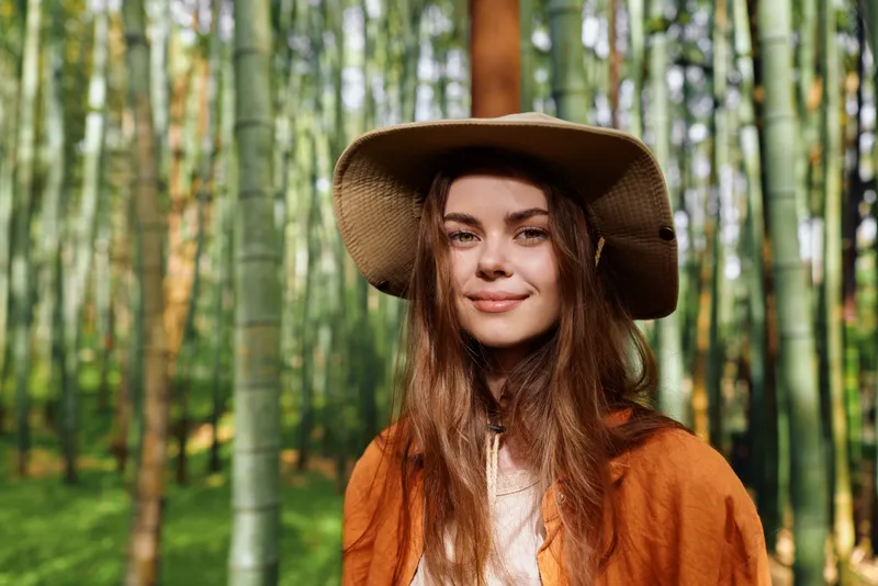 Woman portrait with hat in bamboo forest and nature setting, smiling female model outdoors among green stalks, summer sunlight, casual bohemian style and relaxed expression.