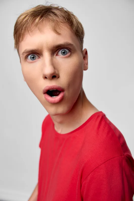 Surprised man in red shirt with wide eyes and open mouth, closeup expression captured in studio, dramatic reaction and curious gaze, intense emotion and astonishment in a vivid candid moment