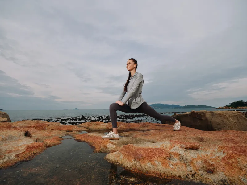 Young athletic woman stretching on rocky beach, wearing sportswear, showcasing a healthy lifestyle and outdoor fitness, under a cloudy sky by the sea