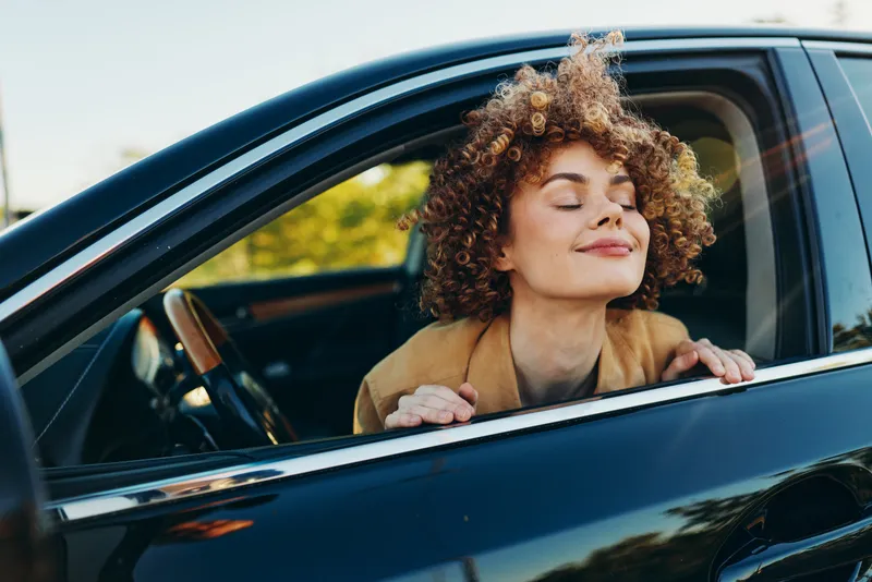 Relaxed woman leans out of a car window with eyes closed, smiling, enjoying a sunny day, casual jacket, retro film color vibe, lifestyle moment