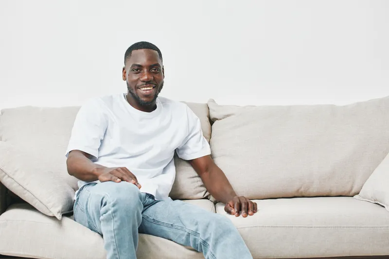 Happy young Black man in casual wear smiling while sitting on a cozy sofa, showcasing comfort and relaxation in a modern living space