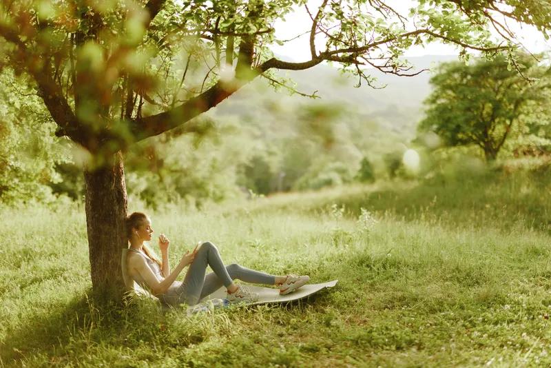 A young woman enjoying a peaceful moment under a tree in a lush green meadow, wearing casual summer clothing and expressing relaxation in a serene outdoor setting