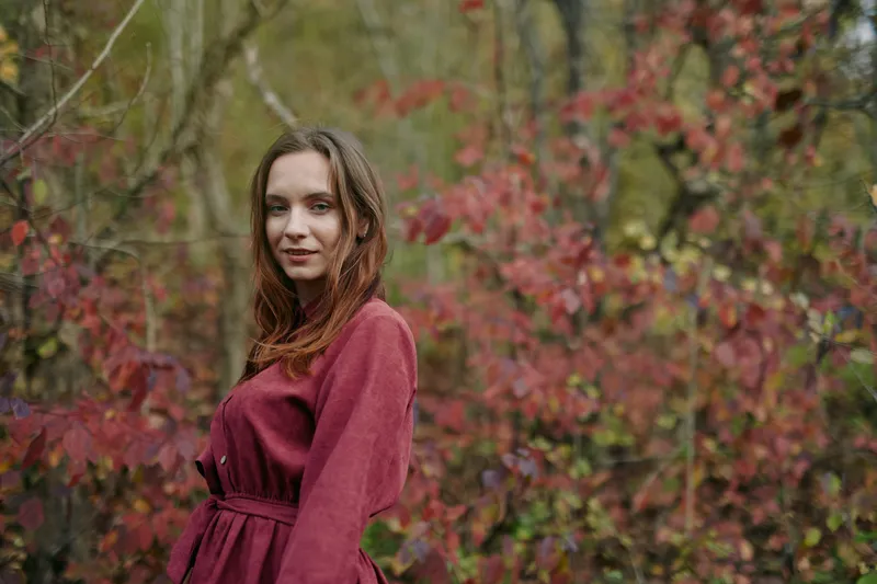 A candid portrait of a young woman in a burgundy dress standing among autumn foliage, conveying authenticity and a natural, unposed mood with soft light.