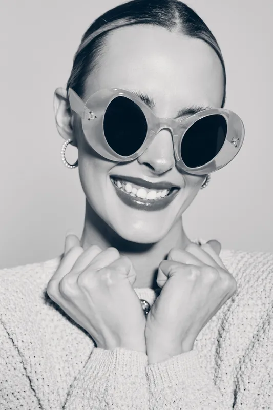 Young white woman with fair skin and slicked-back hair smiles broadly wearing large round sunglasses and hoop earrings, dressed in a textured sweater. Concept of fashion and joy.