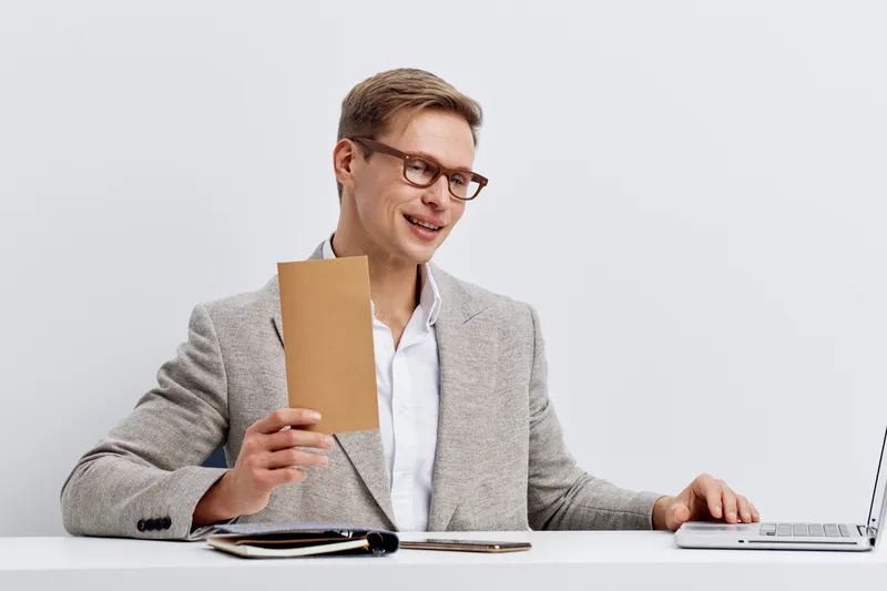 Professional young man with glasses in smart casual attire sitting at white desk with laptop, documents, and notebook, smiling and discussing work in modern office environment. Business, technology, c