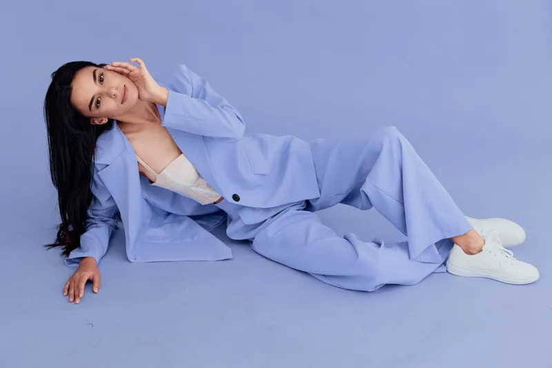 Young woman lying on lavender floor wearing loose blue suit and white sneakers, posing confidently with hand near face against matching pastel background.