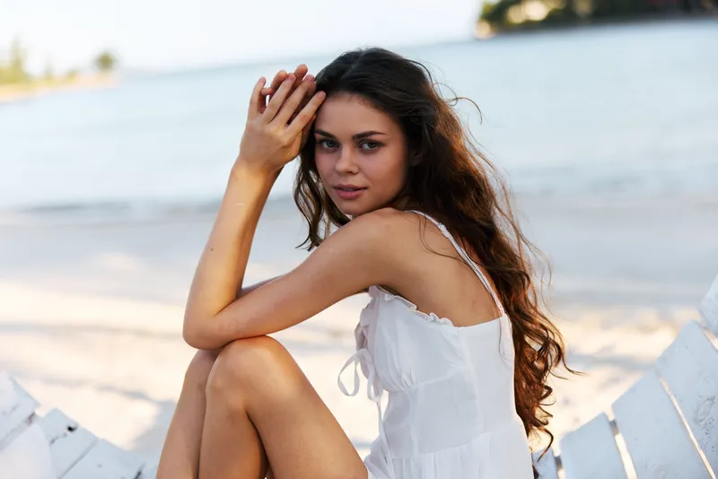 A young woman in a white dress sits on a seaside fence, gazing softly at the camera while warm waves roll ashore under a bright, clear sky.