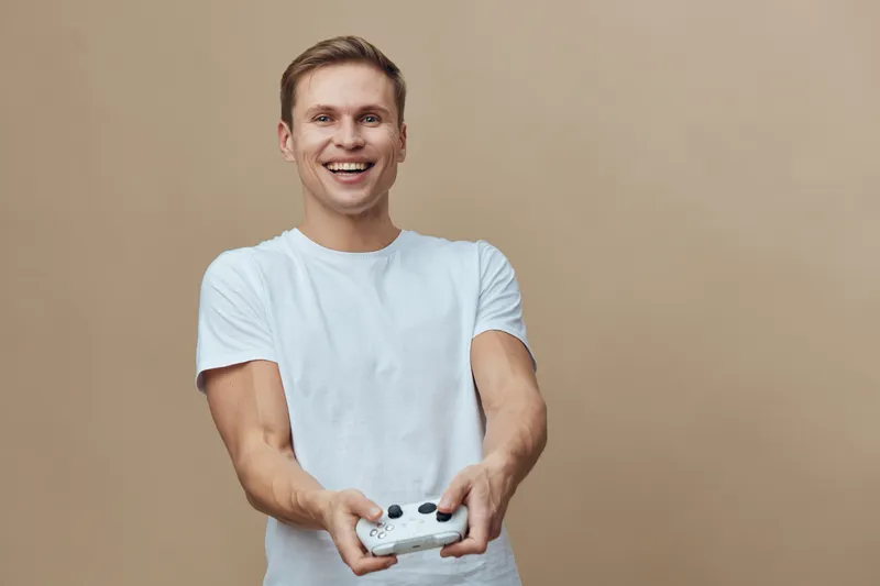 Smiling man holding game controller with both hands, dressed in light blue t-shirt, in studio on beige background, joyful expression, casual style, entertainment and gaming concept.