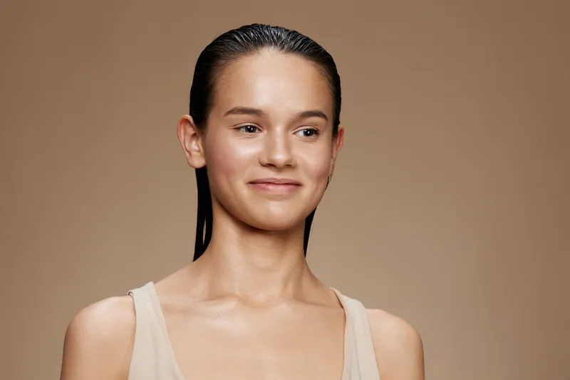 Young Caucasian woman smiling, natural beauty with minimal makeup, wearing a simple beige outfit, showcasing a serene expression against a soft brown background