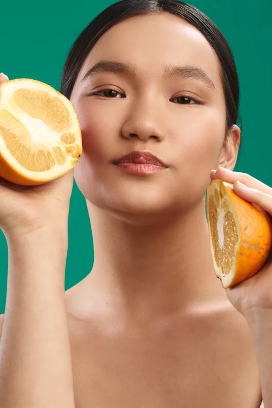 Asian woman holds fresh oranges near her face in a vibrant studio portrait. She gazes calmly at the camera, exuding health, confidence, and a bright, citrusy mood with soft, even lighting.