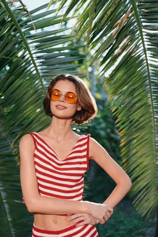 A stylish woman wearing a red striped crop top and round orange sunglasses poses among tropical palm leaves, exuding sunny confidence and carefree charm.