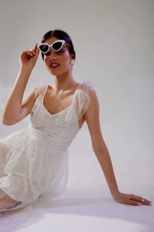 Young woman in a white lace dress wearing stylish sunglasses poses confidently against a neutral background. Concept of woman beauty fashion and elegance.