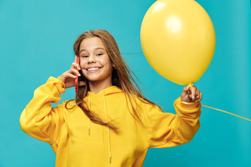 Girl in yellow hoodie holding balloon and talking on smartphone with smile, posing on blue background. Happy kid with long hair wearing casual outfit for leisure and communication.