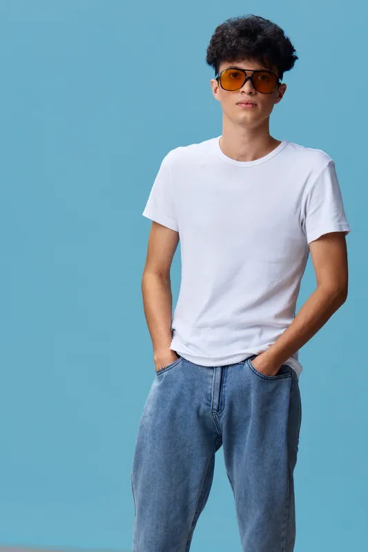 young man wearing sunglasses and casual white t-shirt with hands in pockets standing against blue background with a relaxed confident posture