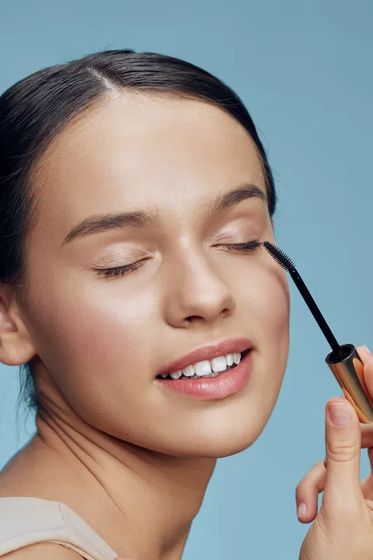 Young woman applying mascara with a joyful expression, showcasing a natural beauty look against a soft blue background, emphasizing self care and confidence in beauty routines