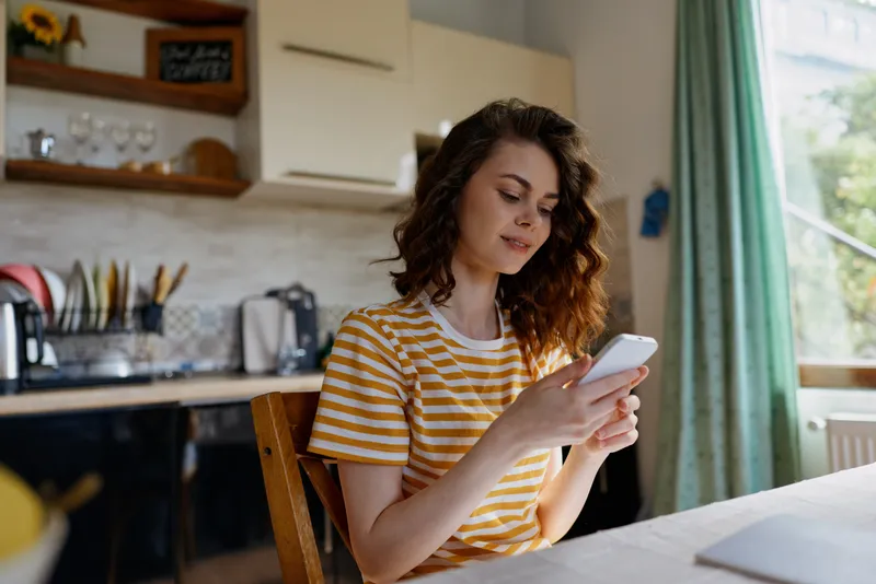 Smiling woman with striped yellow shirt sitting at table, looking at smartphone. Cozy home setting, kitchen in background. Focus on daily routine, self care, and modern living. Soft natural light
