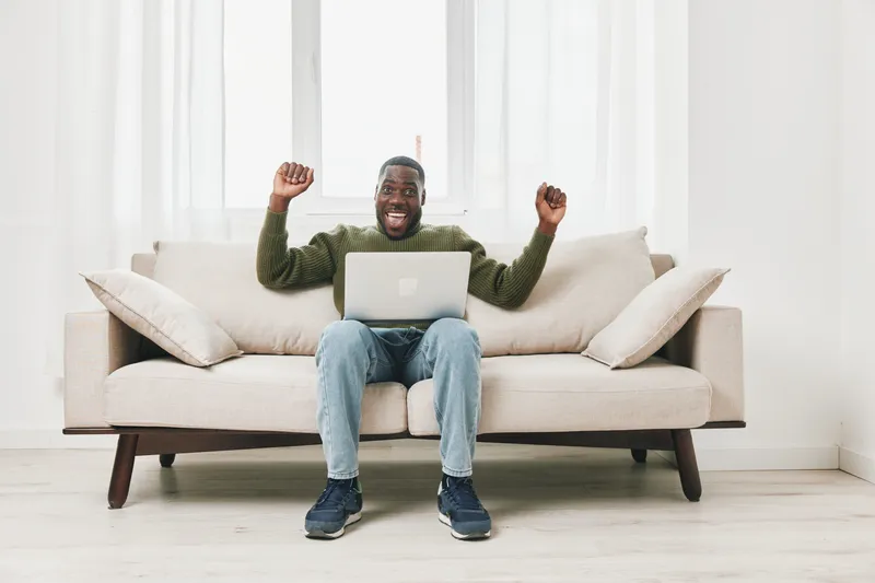 Happy African American man celebrating success while working on a laptop at home, featuring a cozy living room with bright colors and modern decor