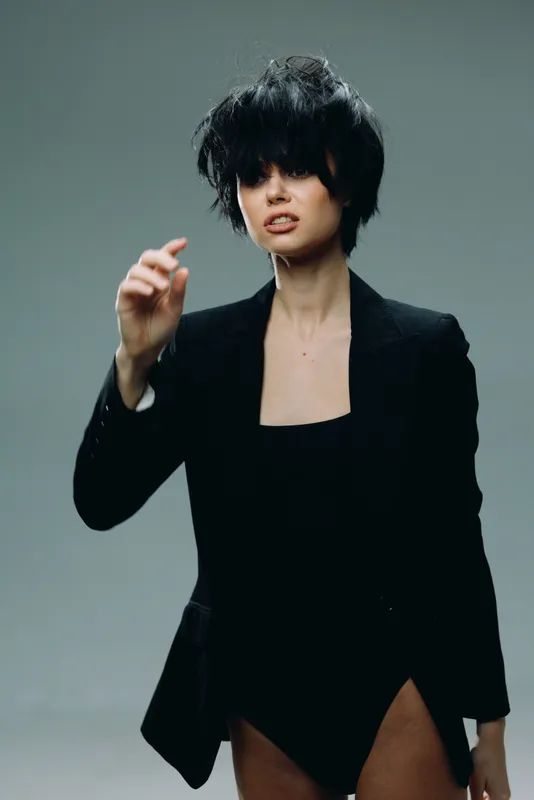 Young woman with dark messy hair wearing black blazer and bodysuit posing against gray background in studio lighting.