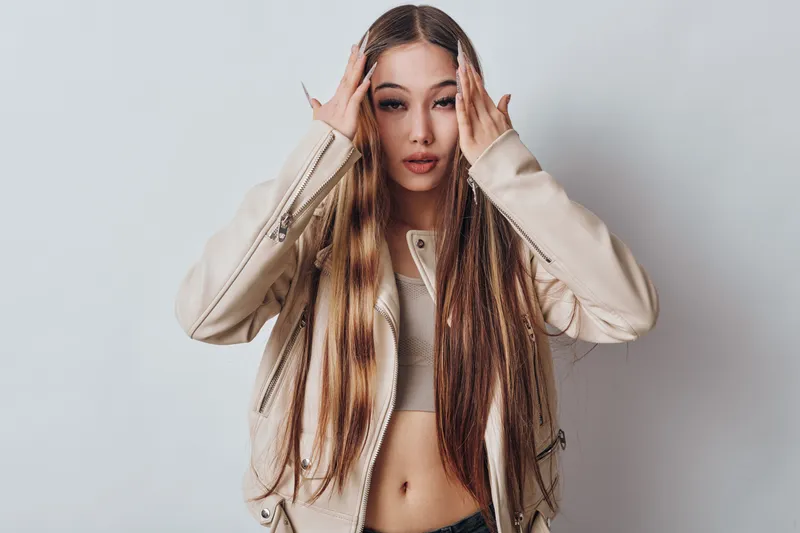 Young woman with long braided hair wearing beige jacket posing in studio. Fashion portrait, casual style with trendy clothing and confident expression on gray background for beauty.