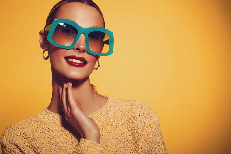 Stylish woman wearing large turquoise sunglasses and yellow sweater poses against orange background. Fashion portrait with bright colors and confident expression for lifestyle concept.