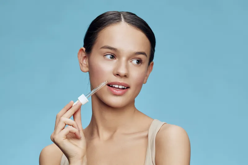 Young woman applying serum with a dropper, showcasing glowing skin and a natural look in a light blue background, representing beauty and skincare