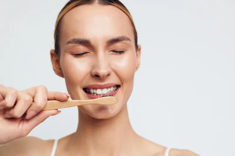 Young woman brushing teeth with bamboo toothbrush, eyes closed, smiling gently, clean fresh skin, white background, personal hygiene, dental care routine