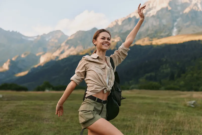 Young woman with a backpack raises her arms in a green valley as distant mountains rise behind, wearing a khaki tied shirt and shorts.