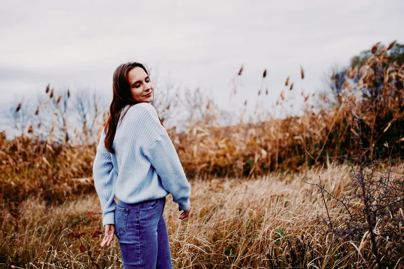 Candid portrait of a young woman walking through tall autumn grasses, conveying gentle authenticity and natural credibility in a relaxed, unposed outdoor moment.