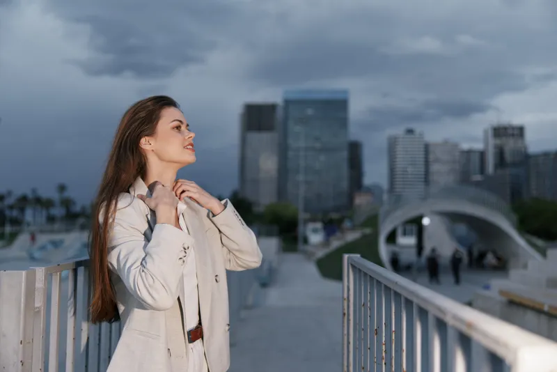 Young business woman in light suit standing confidently outdoors with city skyline and cloudy sky background, expressing calm and determination