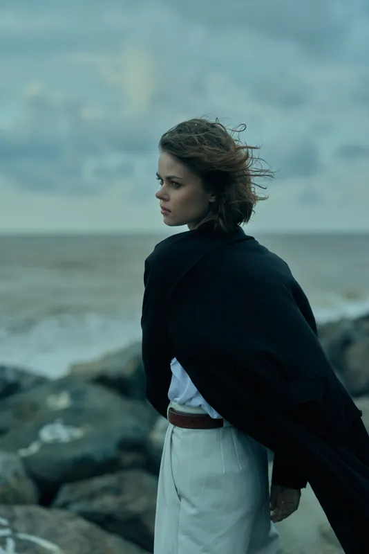 Woman on a rocky coast wrapped in a dark cape gazing toward the sea with a thoughtful expression, wind swept hair, moody clouds, coastal fashion and a solitary wind battered moment