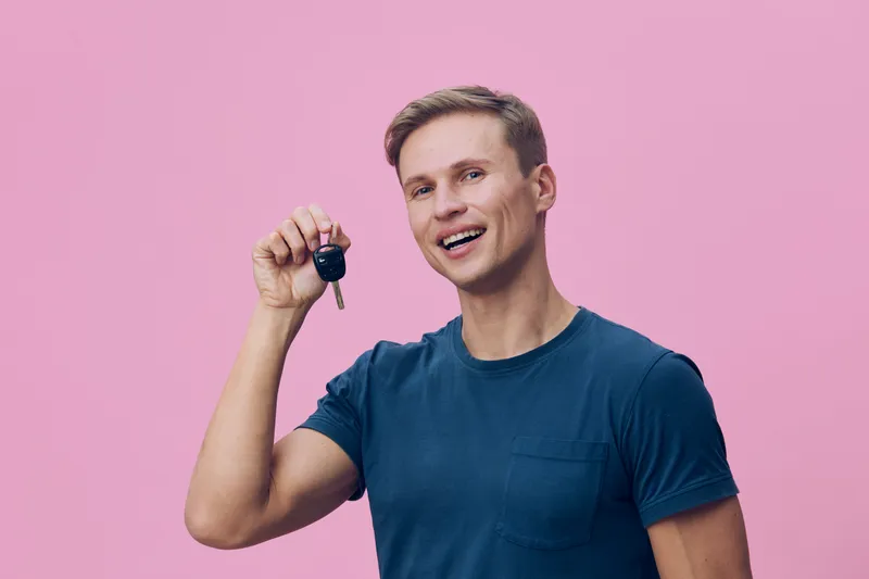 Smiling man wearing casual blue t-shirt holding key with keyring in front of pink background, looking cheerful and confident. Studio portrait of happy person, lifestyle concept, positive emotion, cont