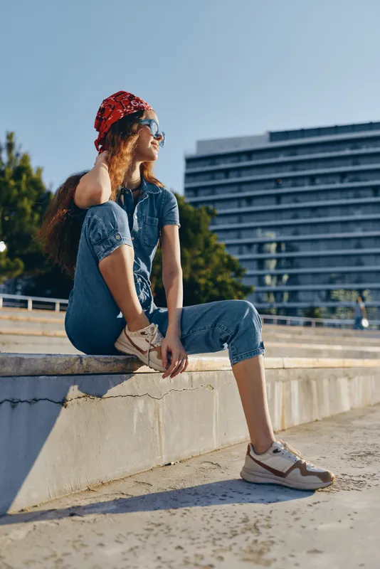 Young woman with red headscarf sitting outdoors on concrete steps in casual denim outfit and sneakers on a sunny day, looking away and enjoying urban environment.