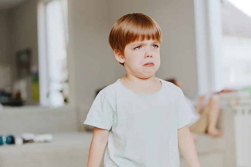 Upset boy standing indoors, expressing sadness and disappointment. The soft colors of the background contribute to the emotional atmosphere of the scene.