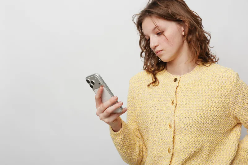 Young woman in yellow sweater looks at smartphone with serious expression on white background. Concept of communication, technology, and thoughtful mood.