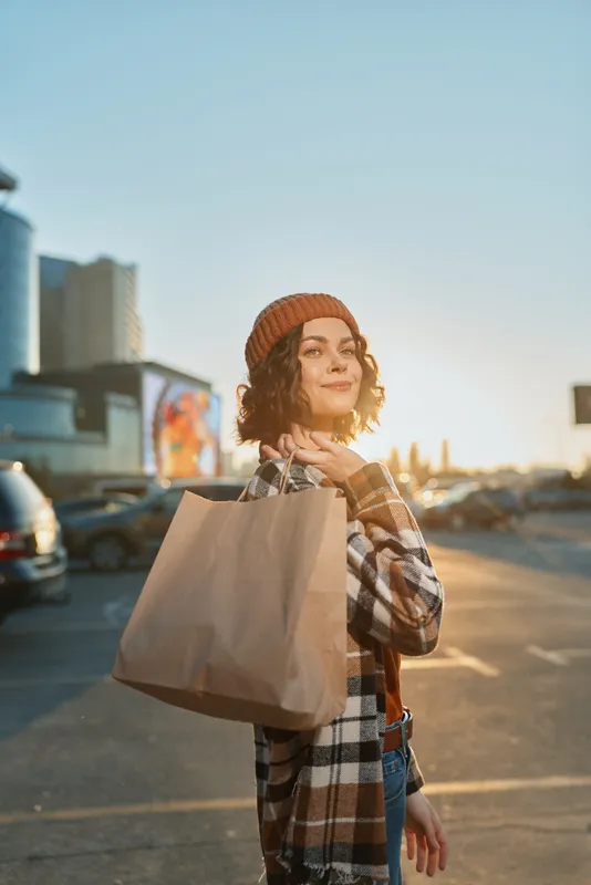 woman shopping bag golden hour candid urban smile: young shopper in beanie carries paper tote in parking lot at sunset, authenticity and candid lifestyle captured with golden hour glow and mindful