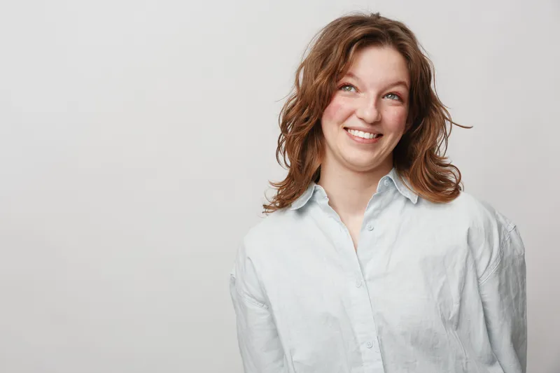 Smiling young woman with curly brown hair wearing white shirt looks up with cheerful expression. Studio portrait on light background. Concept of happiness, positivity, and casual lifestyle.