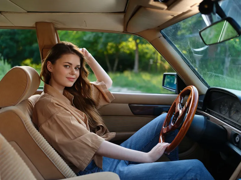 Young woman in casual outfit smiling inside a vintage car, surrounded by greenery, embodying freedom and adventure during a sunny day in nature.