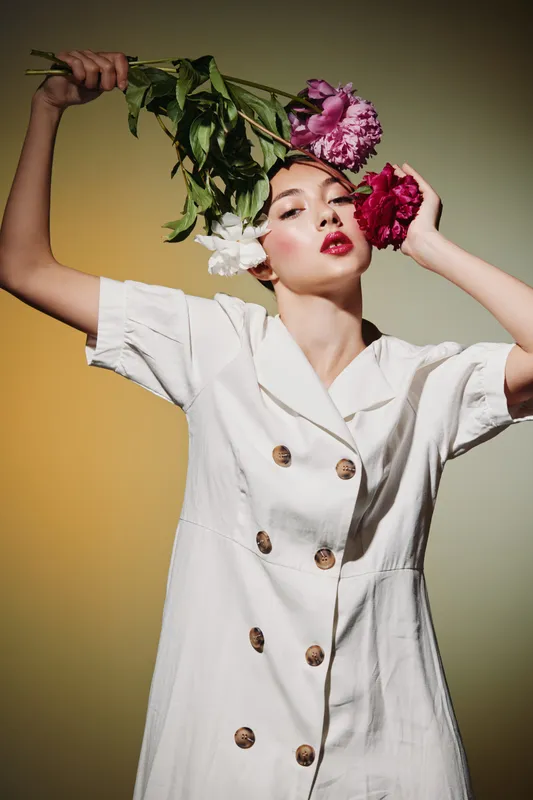 Fashionable woman in white dress posing with colorful flowers near her face on beige background. Creative beauty concept highlighting natural grace and modern style.