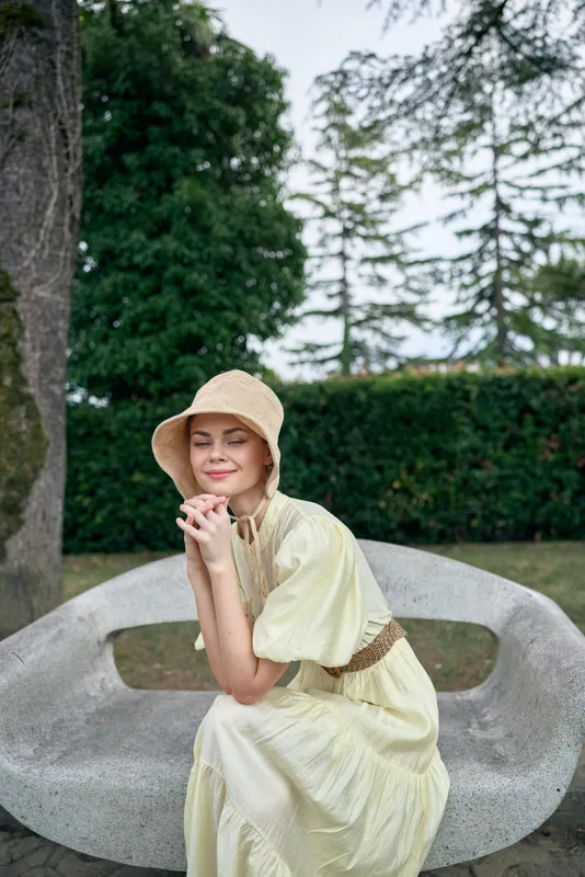 Carefree woman sits on a curved stone bench in a lush garden wearing a light yellow dress and straw hat, posing softly with a gentle smile