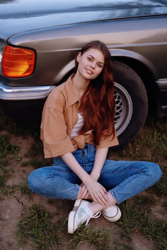 Young woman sitting by a vintage car on a sunny day, wearing a casual outfit with jeans and a shirt, radiating joy and relaxation, perfect for lifestyle concepts.