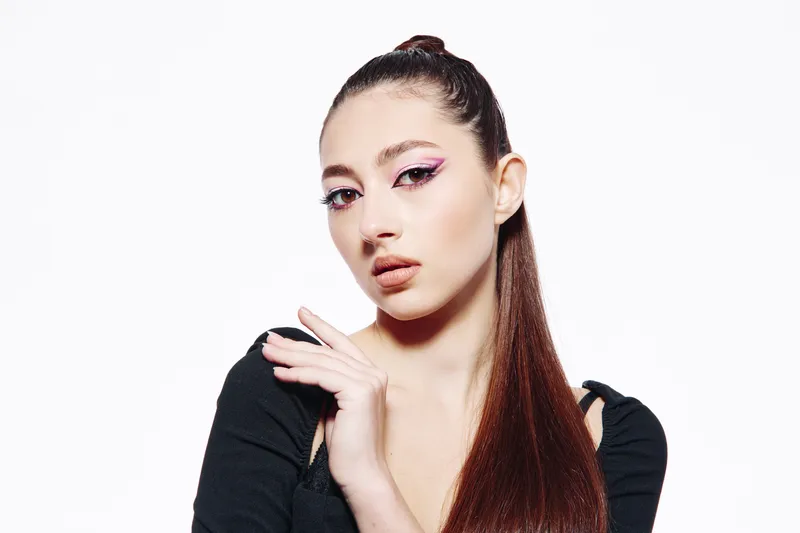 Young woman with creative bright eye makeup poses with hand near face. Long sleek hair is styled in a top knot. White background highlights bold fashion and beauty concept.