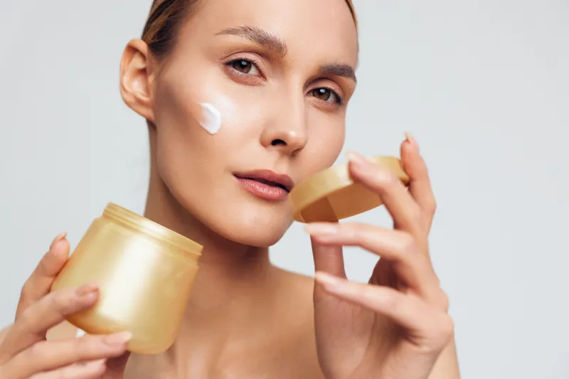 A young woman applies moisturizing cream on her cheek using her finger while holding an open jar of skincare product. The clean background highlights natural beauty and self-care routine.