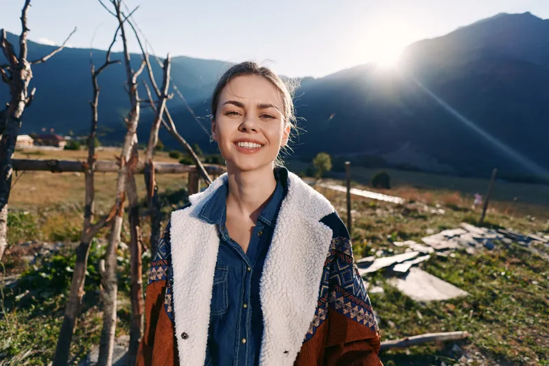 Woman smile mountains countryside portrait sunlight outdoors traveler jacket female in meadow, casual denim shirt and shearling coat, backlit happy headshot with rustic fence and scenic landscape.
